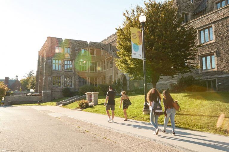 Photo of four students walking in front of Hamilton Hall in summer with the sun shining down on them.